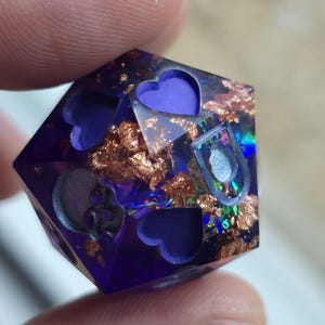 May include: A handmade, multi-faceted, purple resin die with embedded gold flakes, hearts, and a skull. The die is held between two fingers, showcasing its intricate details and unique design. The die is a perfect gift for tabletop gamers.