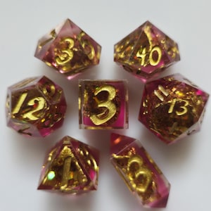 May include: Set of seven polyhedral dice with a pink and gold color scheme. The dice are transparent with gold-colored numbers and glittery gold accents. The dice are arranged in a circular pattern.