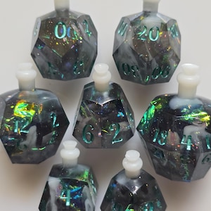May include: Seven polyhedral dice shaped like potion bottles. The dice are dark gray with iridescent glitter and teal numbers. Each bottle has a white stopper. The dice are arranged on a white surface.