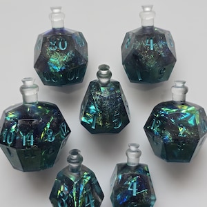 Puede incluir: Una colección de siete botellas de vidrio en forma de dados con tapones transparentes. Las botellas son de color verde azulado oscuro con remolinos iridiscentes azules y verdes. Cada botella tiene un número diferente grabado en su superficie, del 0 al 14. Las botellas están dispuestas sobre un fondo blanco.