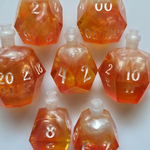 Puede incluir: Siete dados de resina naranja y amarilla con números blancos y una parte superior en forma de botella. Los dados están numerados con varios valores. Los dados están dispuestos sobre una superficie blanca.