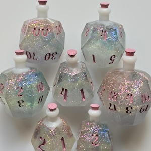 May include: Seven glittery dice-shaped bottles with pink numbers and stoppers. The bottles are clear with iridescent glitter, and the numbers are in a pink font. The bottles are arranged in a circular pattern.