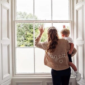 May include: A tan crochet sweater with the name "Pagett" embroidered on the back in white script, along with a small heart. The sweater is worn by a person holding a child near a window. The window has white trim.