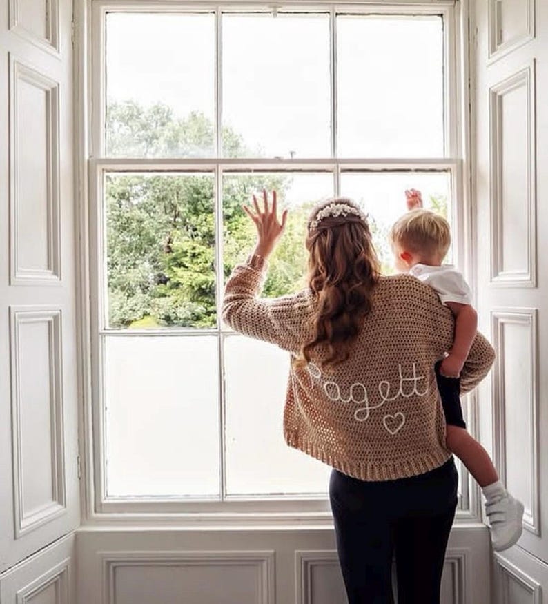May include: A tan crochet sweater with the name "Pagett" embroidered on the back in white script, along with a small heart. The sweater is worn by a person holding a child near a window. The window has white trim.