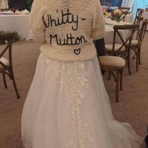 May include: A cream-colored, crocheted sweater with the words "Whitty-Mutton" embroidered in black script, worn over a white wedding dress. The dress features delicate floral embroidery. A heart is embroidered below the name.