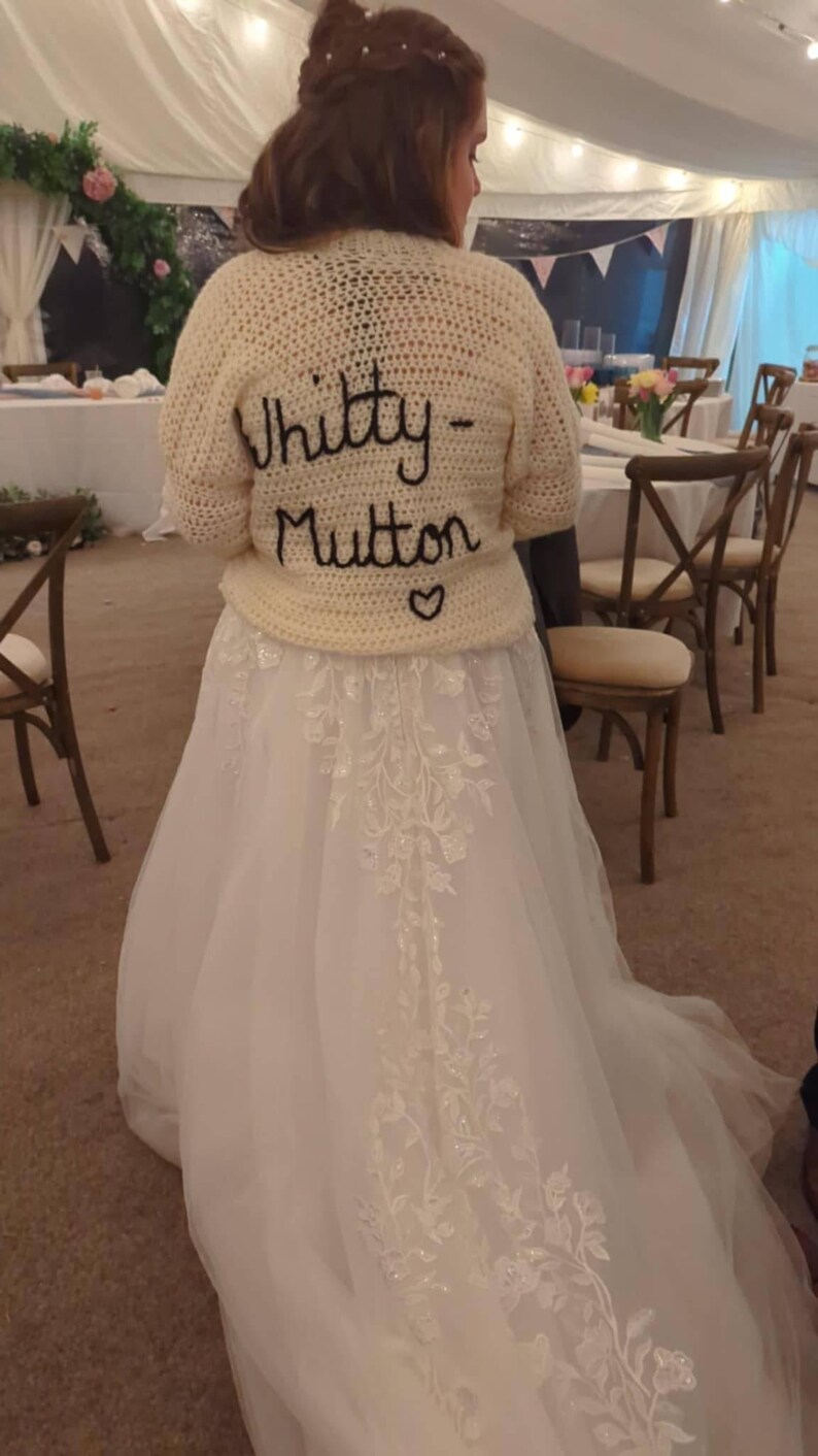 May include: A cream-colored, crocheted sweater with the words "Whitty-Mutton" embroidered in black script, worn over a white wedding dress. The dress features delicate floral embroidery. A heart is embroidered below the name.