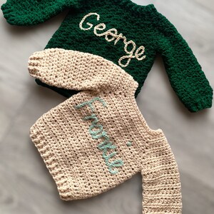 May include: Two long-sleeved crochet sweaters. The top sweater is dark green with the name George embroidered in cream yarn. The bottom sweater is beige with the name Frankie embroidered in light blue yarn. Both feature a textured stitch pattern.