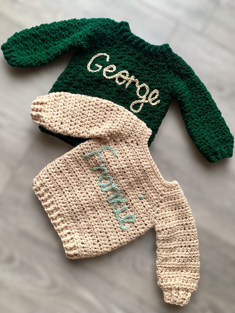 May include: Two long-sleeved crochet sweaters. The top sweater is dark green with the name George embroidered in cream yarn. The bottom sweater is beige with the name Frankie embroidered in light blue yarn. Both feature a textured stitch pattern.