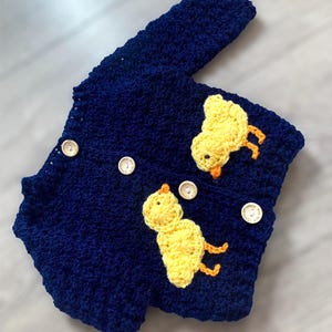 May include: A dark blue crocheted cardigan featuring long sleeves and four light-colored wooden buttons. Two yellow crocheted chick appliqu&eacute;s with orange beaks and feet are attached to the front of the garment, one on each side of the button placket.