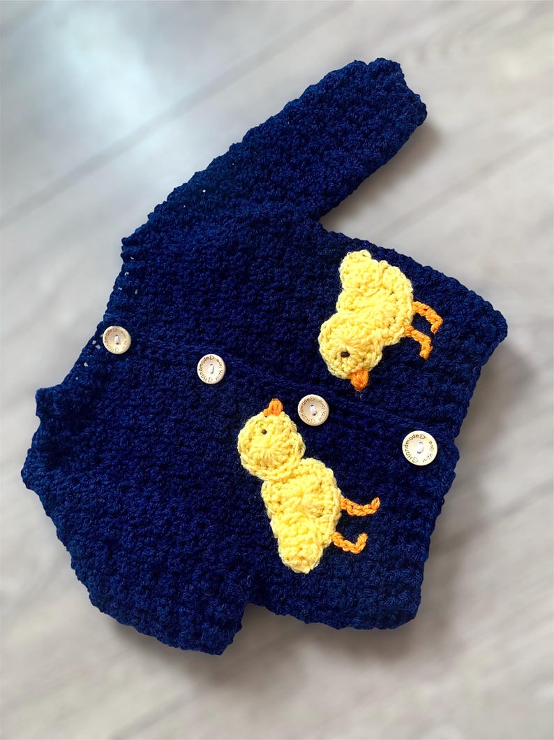 May include: A dark blue crocheted cardigan featuring long sleeves and four light-colored wooden buttons. Two yellow crocheted chick appliqu&eacute;s with orange beaks and feet are attached to the front of the garment, one on each side of the button placket.