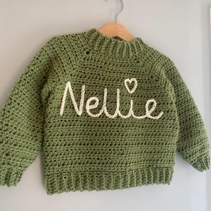 May include: A sage green long-sleeve crochet sweater featuring a ribbed neckline, cuffs, and hem. The front is embroidered with the name Nellie in cream-colored yarn, accompanied by a small heart symbol positioned above the letter i.