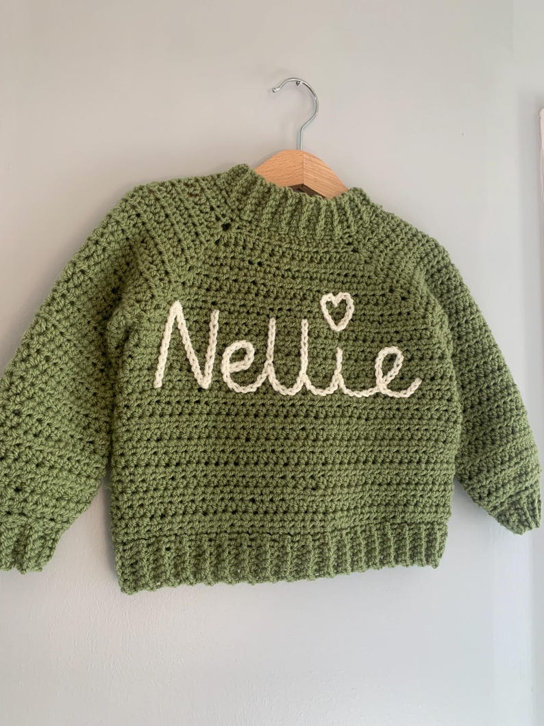 May include: A sage green long-sleeve crochet sweater featuring a ribbed neckline, cuffs, and hem. The front is embroidered with the name Nellie in cream-colored yarn, accompanied by a small heart symbol positioned above the letter i.