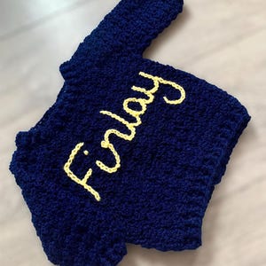 May include: A dark blue crocheted garment featuring the name Finlay stitched in yellow yarn across the front. The item has a textured, looped surface and a short-sleeved silhouette, showcasing a handmade construction with visible yarn stitches.