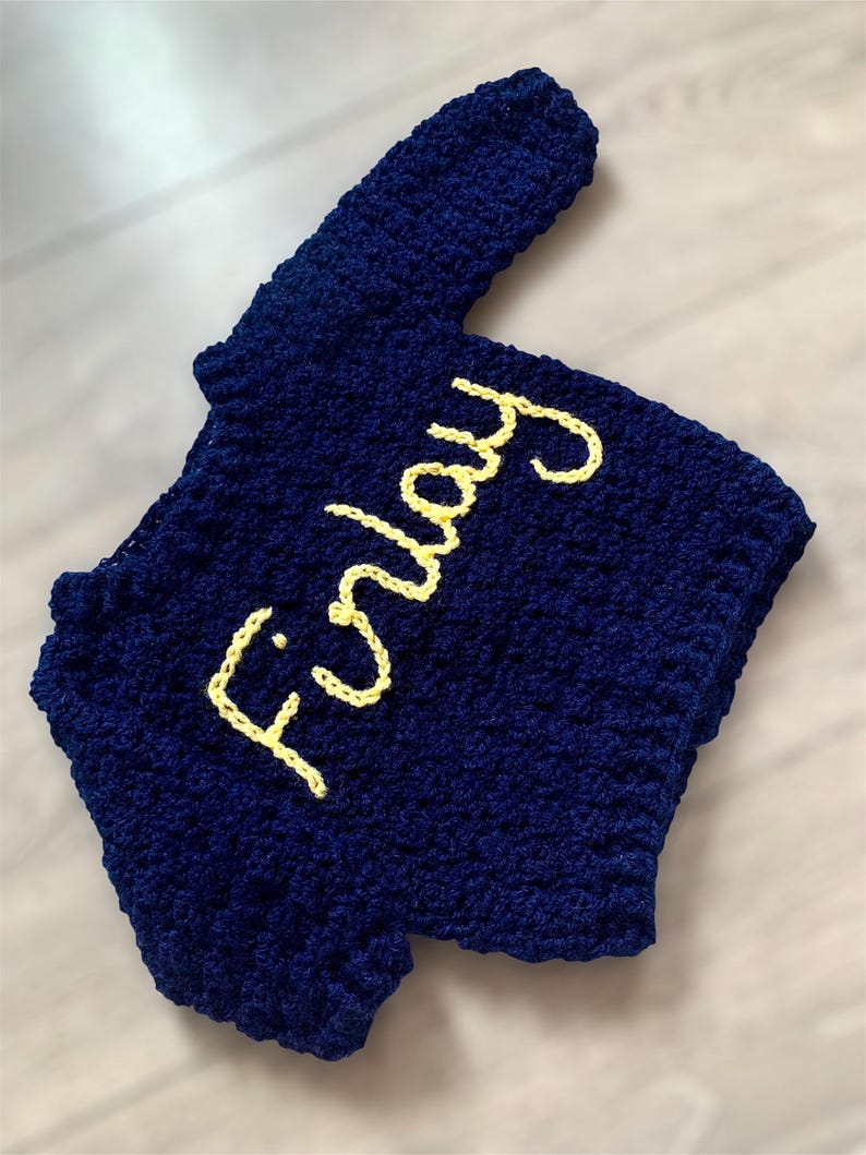 May include: A dark blue crocheted garment featuring the name Finlay stitched in yellow yarn across the front. The item has a textured, looped surface and a short-sleeved silhouette, showcasing a handmade construction with visible yarn stitches.