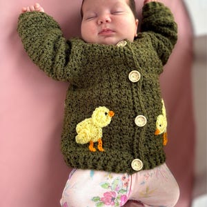 May include: An olive green crocheted long-sleeve cardigan featuring three light-colored wooden buttons with the text handmade with love and two yellow crocheted chick appliqu&eacute;s with orange beaks and feet, worn over light pink floral patterned pants.