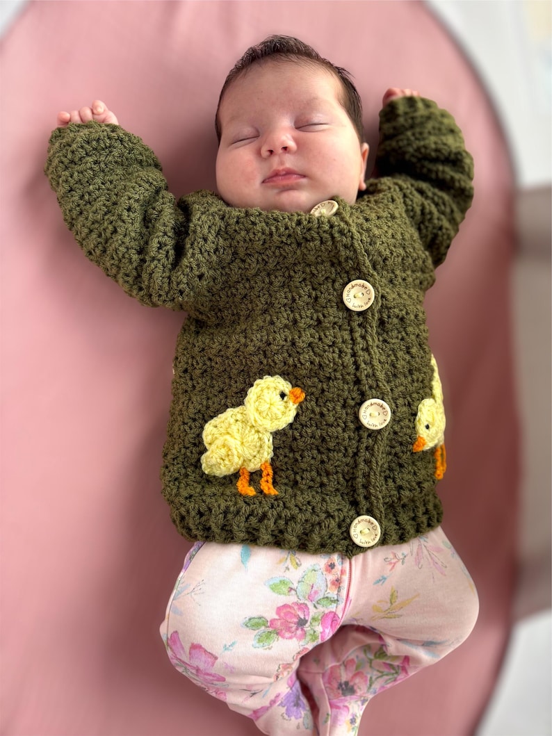 May include: An olive green crocheted long-sleeve cardigan featuring three light-colored wooden buttons with the text handmade with love and two yellow crocheted chick appliqu&eacute;s with orange beaks and feet, worn over light pink floral patterned pants.