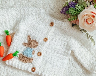 May include: A white crocheted long-sleeve cardigan featuring a brown and light blue bunny appliqué on the front. The garment includes four round wooden buttons along the center placket and a textured stitch pattern throughout the fabric.