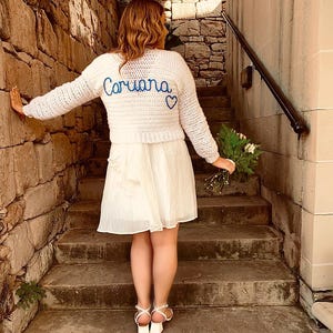 May include: A white crochet sweater with the name "Coriana" in blue script and a blue heart. The sweater is paired with a white dress and white sandals. The person is standing on stone steps, holding a bouquet of flowers.