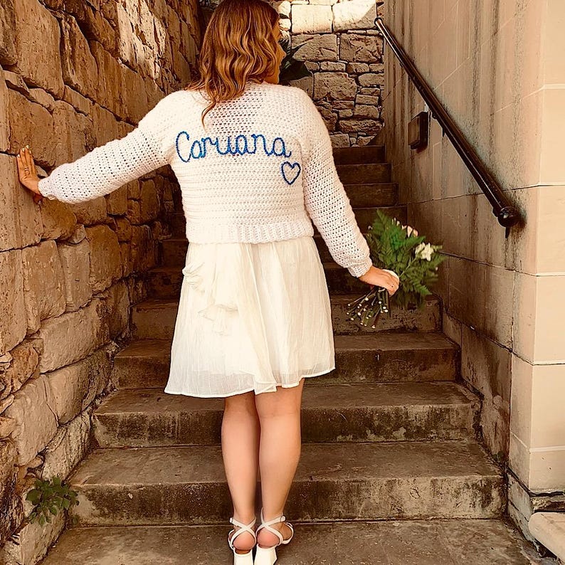 May include: A white crochet sweater with the name "Coriana" in blue script and a blue heart. The sweater is paired with a white dress and white sandals. The person is standing on stone steps, holding a bouquet of flowers.