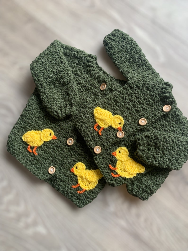 May include: Two dark olive green crocheted cardigans featuring long sleeves and round wooden buttons. Each garment is decorated with bright yellow crocheted ducklings that have orange beaks and feet, stitched onto the textured yarn surface.