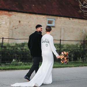 May include: A bride in a white wedding dress with a long train and a bouquet of flowers walks with a person in a black suit. The dress has the word "lalite" and a heart symbol on the back.