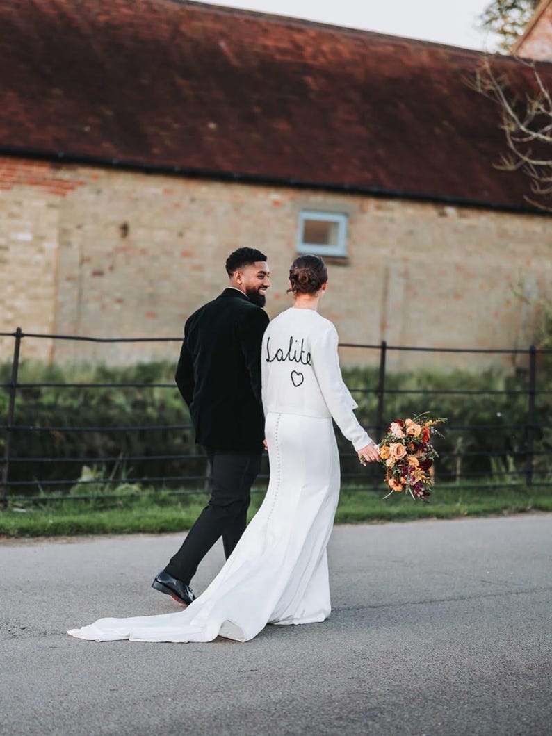 May include: A bride in a white wedding dress with a long train and a bouquet of flowers walks with a person in a black suit. The dress has the word "lalite" and a heart symbol on the back.