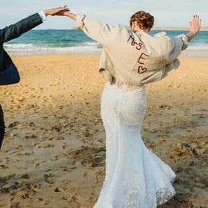 May include: A bride wearing a white lace wedding dress and a tan jacket with "Mrs E" embroidered on the back, with two hearts. The couple is on a sandy beach with the ocean in the background.