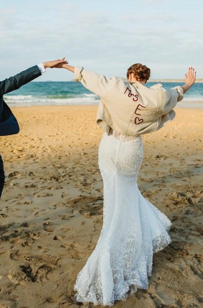 May include: A bride wearing a white lace wedding dress and a tan jacket with "Mrs E" embroidered on the back, with two hearts. The couple is on a sandy beach with the ocean in the background.