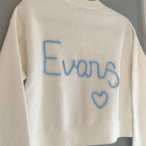 May include: Cream-colored sweater with long sleeves. The word "Evans" is embroidered in light blue thread across the back, along with a blue heart. The sweater is hanging on a clothes hanger.