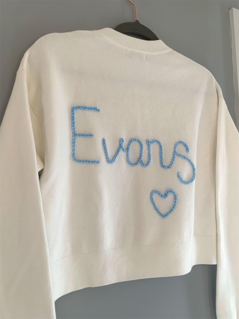 May include: Cream-colored sweater with long sleeves. The word "Evans" is embroidered in light blue thread across the back, along with a blue heart. The sweater is hanging on a clothes hanger.