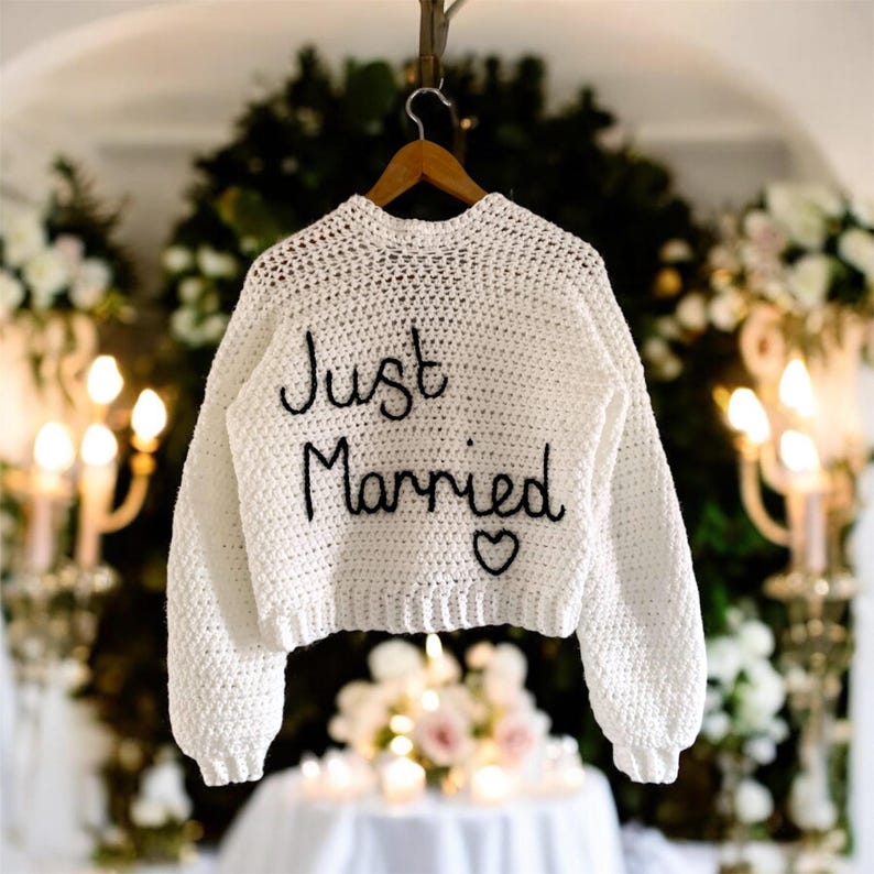 May include: White crochet sweater with the words "Just Married" embroidered in black script. A small black heart is embroidered below the text. The sweater is hanging on a wooden hanger.