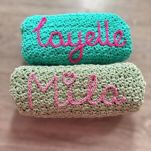 May include: Two folded crochet blankets stacked vertically. The top blanket is turquoise with the name Layelle embroidered in pink yarn. The bottom blanket is sage green with the name Mila and a small heart embroidered in pink yarn.