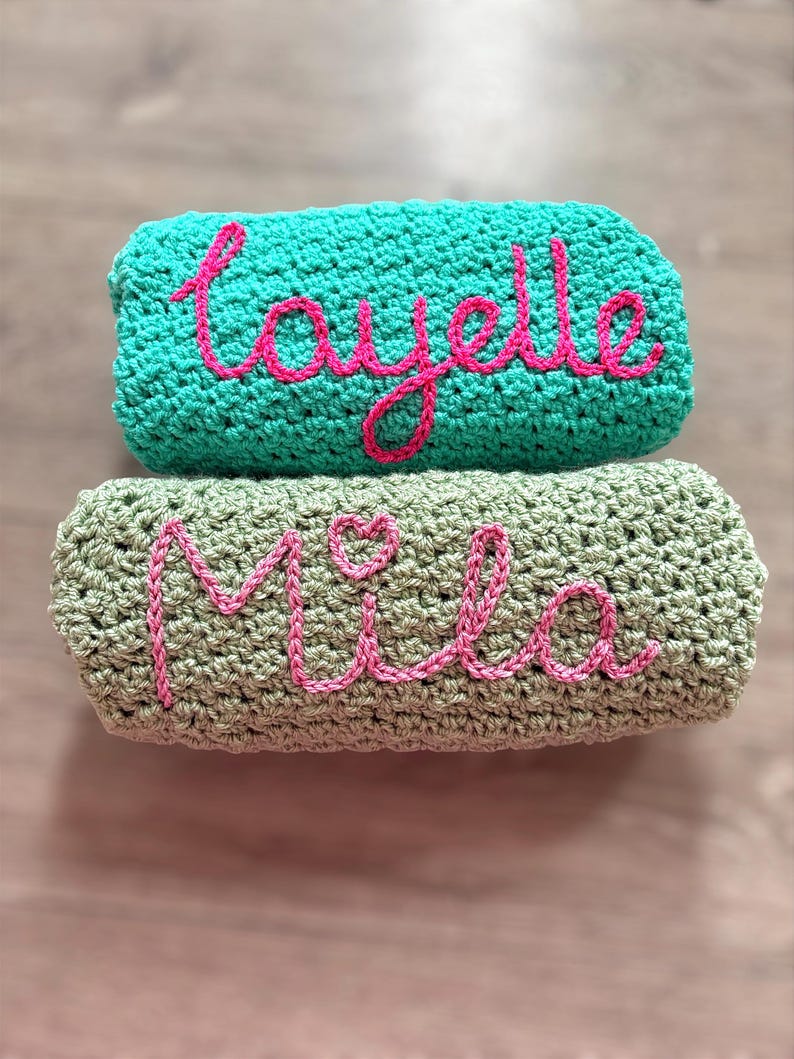 May include: Two folded crochet blankets stacked vertically. The top blanket is turquoise with the name Layelle embroidered in pink yarn. The bottom blanket is sage green with the name Mila and a small heart embroidered in pink yarn.
