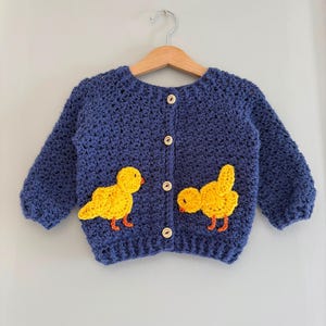 May include: A navy blue long-sleeve crochet cardigan featuring four light-colored wooden buttons down the center. Two bright yellow crocheted chick appliqu&eacute;s with orange beaks and feet are attached to the front of the garment on either side.
