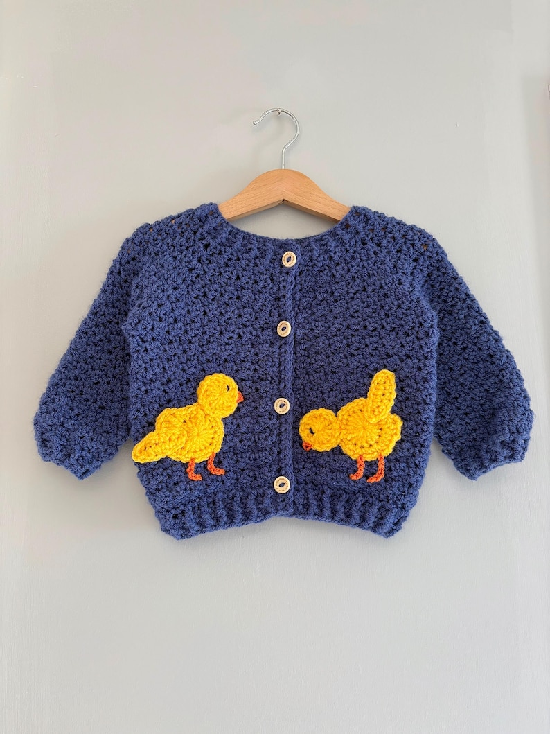 May include: A navy blue long-sleeve crochet cardigan featuring four light-colored wooden buttons down the center. Two bright yellow crocheted chick appliqu&eacute;s with orange beaks and feet are attached to the front of the garment on either side.