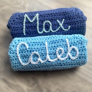 May include: Two stacked rectangular crochet blankets. The top blanket is dark navy blue with the name Max embroidered in light mint green yarn. The bottom blanket is a lighter sky blue with the name Caleb embroidered in white yarn.