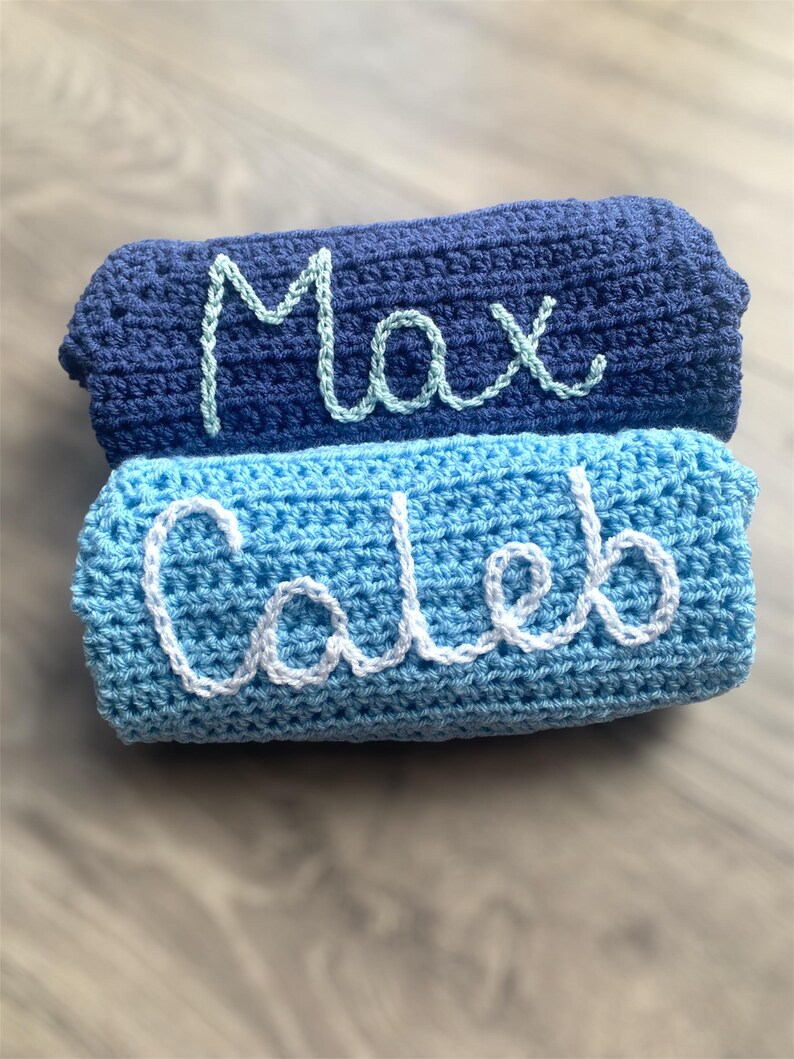 May include: Two stacked rectangular crochet blankets. The top blanket is dark navy blue with the name Max embroidered in light mint green yarn. The bottom blanket is a lighter sky blue with the name Caleb embroidered in white yarn.