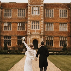 May include: A couple walks away from the camera on a gravel path, holding hands. The person in front wears a white jacket with the word "Cox" on the back. A large brick building with many windows is in the background.