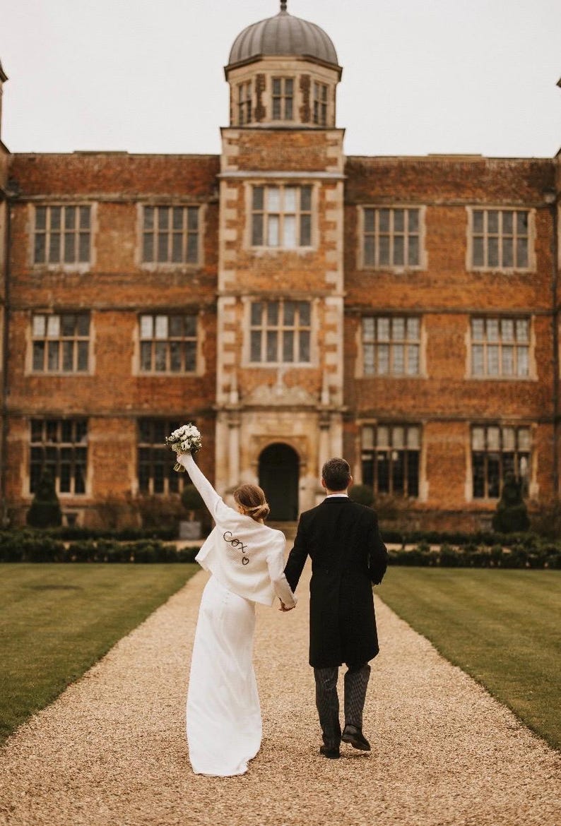 May include: A couple walks away from the camera on a gravel path, holding hands. The person in front wears a white jacket with the word "Cox" on the back. A large brick building with many windows is in the background.
