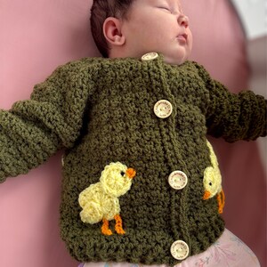 May include: A long-sleeved olive green crochet cardigan featuring two yellow chick appliqu&eacute;s with orange beaks and feet. The garment fastens with four round wooden buttons, each engraved with the text "handmade with love" and a heart icon.