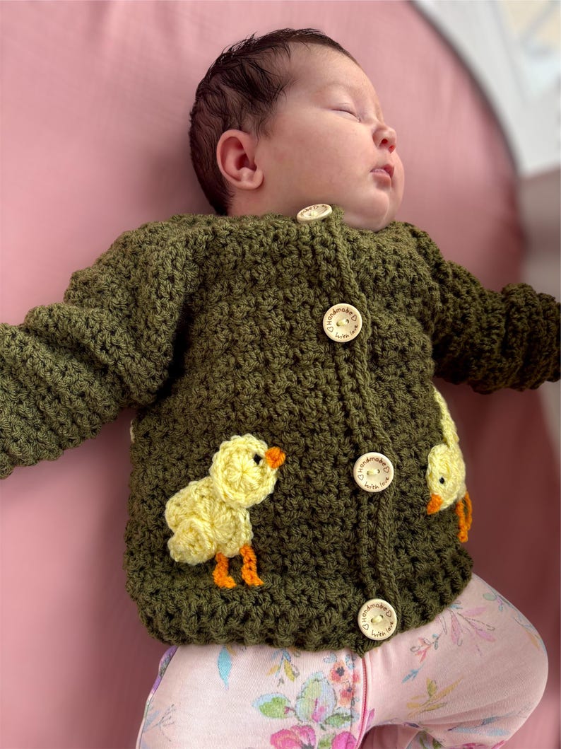 May include: A long-sleeved olive green crochet cardigan featuring two yellow chick appliqu&eacute;s with orange beaks and feet. The garment fastens with four round wooden buttons, each engraved with the text "handmade with love" and a heart icon.