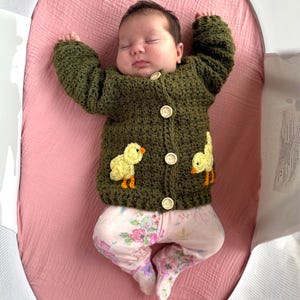 May include: A dark olive green crocheted long-sleeve cardigan featuring four light-colored wooden buttons down the center and two yellow chick appliques on the lower front panels, worn over light-colored pants with a pink and purple floral print.