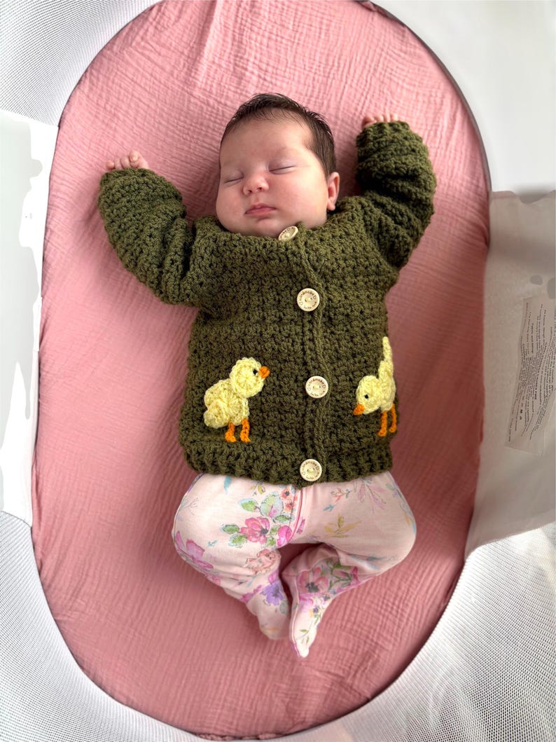 May include: A dark olive green crocheted long-sleeve cardigan featuring four light-colored wooden buttons down the center and two yellow chick appliques on the lower front panels, worn over light-colored pants with a pink and purple floral print.
