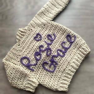 May include: A beige long-sleeved crochet sweater featuring the name Rosie Grace embroidered in purple yarn across the front, accompanied by a small purple heart symbol. The garment has a ribbed neckline, cuffs, and hem with a textured stitch pattern.