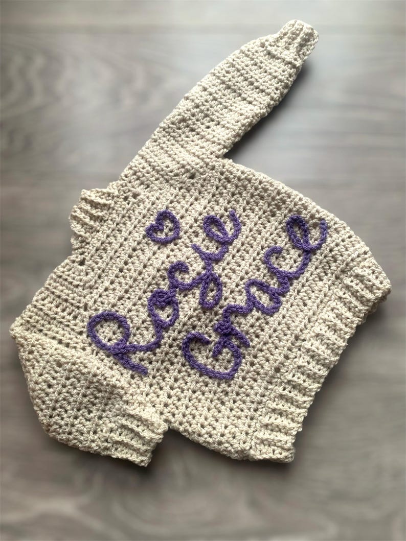 May include: A beige long-sleeved crochet sweater featuring the name Rosie Grace embroidered in purple yarn across the front, accompanied by a small purple heart symbol. The garment has a ribbed neckline, cuffs, and hem with a textured stitch pattern.