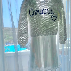 May include: A white crochet sweater with long sleeves and the name "Caruana" embroidered in navy blue, along with a navy blue heart. The sweater is paired with a white dress, hanging in front of a window.