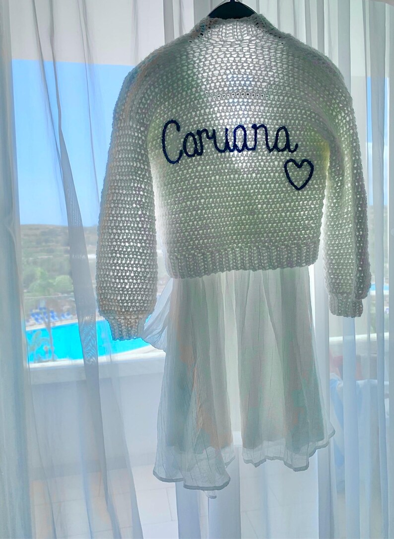 May include: A white crochet sweater with long sleeves and the name "Caruana" embroidered in navy blue, along with a navy blue heart. The sweater is paired with a white dress, hanging in front of a window.