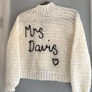 May include: Cream-colored, hand-crocheted sweater with long sleeves and a high neckline. The words "Mrs Davis" are embroidered in black script across the front, with a small heart symbol below. The sweater is hanging on a hanger.