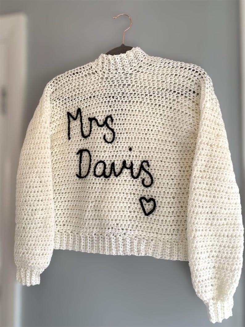 May include: Cream-colored, hand-crocheted sweater with long sleeves and a high neckline. The words "Mrs Davis" are embroidered in black script across the front, with a small heart symbol below. The sweater is hanging on a hanger.