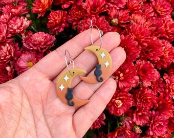 May include: A pair of hoop earrings featuring crescent moon charms in a gradient of orange and yellow. Each moon charm is decorated with a small black cat silhouette and two white four-pointed star shapes, suspended from thin silver-toned metal hoops.
